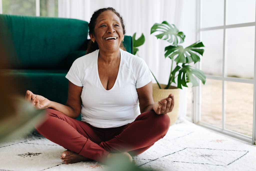 Black elderly woman meditating in lotus position at home, wearing a happy smile. Retired senior prioritizing her mental health and wellbeing, finding peace and serenity in her yoga practice. practicing mindfulness in seniors